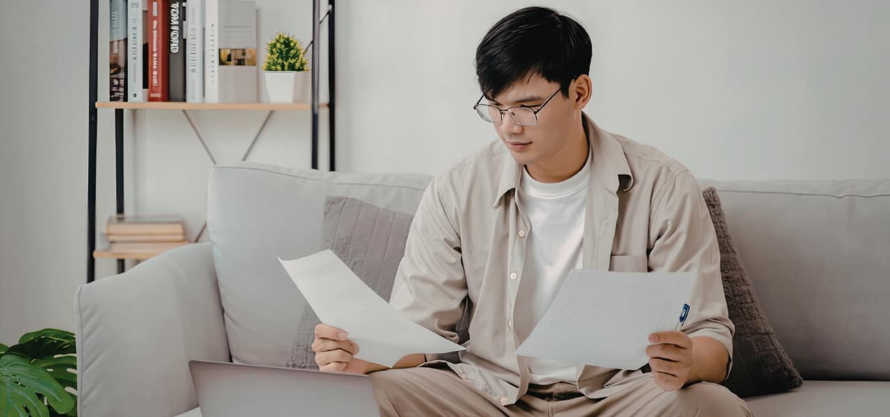 Person sitting on a couch at home reviewing documents related to an insurance claim while using a laptop, appearing to carefully compare total loss settlement paperwork.