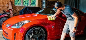 A man washing a red sports car by hand inside a garage, cleaning the exterior with a sponge and soap to maintain the vehicle’s appearance.