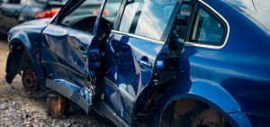 A blue sedan with heavy side impact damage and missing wheels sitting in a salvage yard