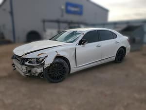 A white luxury sedan with significant front-end damage, including a missing bumper, exposed components, and dents along the front left side, parked in an outdoor lot near an industrial building.